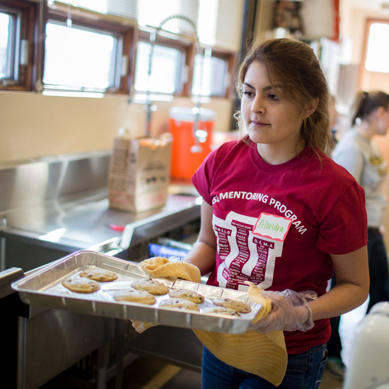 Female in commercial kitchen carrying tray of cookies.